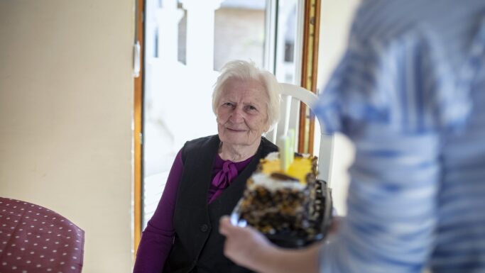 En ældre kvinde med hvidt hår sidder ved et bord og smiler, mens nogen rækker hende et stykke kage med frugt på toppen. Sollyset strømmer ind gennem vinduet i baggrunden og kaster et blidt skær over den 90-årige kvindes glade ansigtsudtryk.