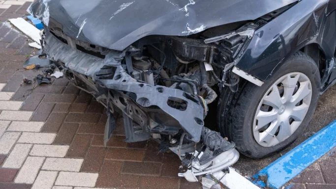 En sort bil med alvorlige skader på fronten står parkeret på en parkeringsplads sammen med andre biler. Kofangeren og forlygten mangler, hvilket blotlægger indvendige dele, hvilket tyder på, at ulykken muligvis involverede en fodgænger eller måske en fejl med pedalerne. Den hvidstribede fortovsbelægning kan ses under og omkring køretøjet.