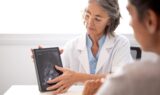 En læge i hvid kittel viser en patient et scanningsbillede på en tablet og forklarer resultaterne af patientens kræftforløb under en konsultation i et lyst rum, mens de drøfter, hvordan patientens helbred kan forbedres.