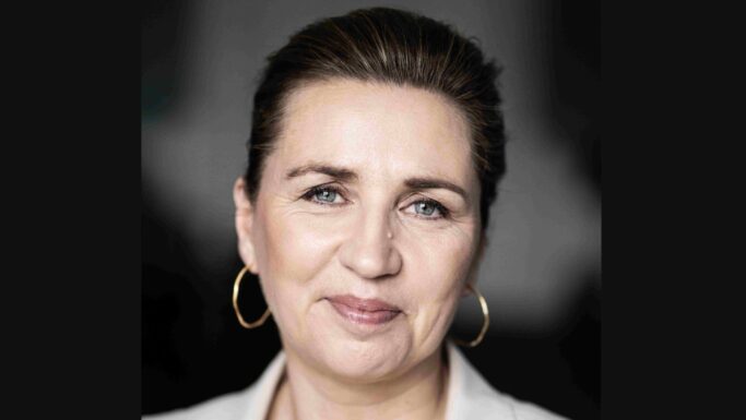 En kvinde med lys hud, blå øjne og mørkt hår, der er sat op, iført guldringe i ørerne og en lys bluse, smiler blidt foran en sløret, mørk baggrund – et billede, der indfanger den rolige selvsikkerhed hos en person, der taler om madpriser og lavere mødre med en statsministers autoritet.