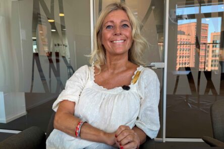 En blond kvinde i en hvid bluse sidder smilende i et moderne kontor med glasvægge, og bag hende kan man gennem vinduerne se byens bygninger; hendes pengepung er klar til at blive fyldt op med nye muligheder.