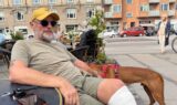 En ældre mand med en gul hat og et forbandet knæ sidder ved et bord på en udendørs café med en telefon og nogle nøgler. Hans trofaste brune hund står ved siden af ham, mens han nyder byens gadeliv og de omkringliggende bygninger, tilsyneladende uanfægtet af enhver skræksygdom.