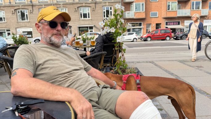 En ældre mand med en gul hat og et forbandet knæ sidder ved et bord på en udendørs café med en telefon og nogle nøgler. Hans trofaste brune hund står ved siden af ham, mens han nyder byens gadeliv og de omkringliggende bygninger, tilsyneladende uanfægtet af enhver skræksygdom.