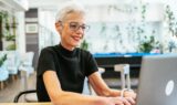 En ældre kvinde med kort, gråt hår og briller smiler, mens hun arbejder på en bærbar computer i et lyst, moderne kontor fyldt med planter – et glimrende eksempel på, hvordan arbejdspladser bliver mere inkluderende for seniorer.
