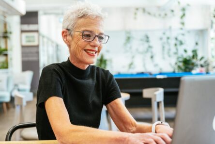 En ældre kvinde med kort, gråt hår og briller smiler, mens hun arbejder på en bærbar computer i et lyst, moderne kontor fyldt med planter – et glimrende eksempel på, hvordan arbejdspladser bliver mere inkluderende for seniorer.
