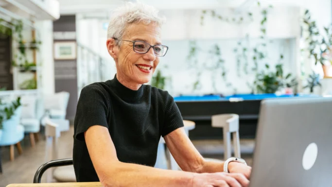 En ældre kvinde med kort, gråt hår og briller smiler, mens hun arbejder på en bærbar computer i et lyst, moderne kontor fyldt med planter – et glimrende eksempel på, hvordan arbejdspladser bliver mere inkluderende for seniorer.