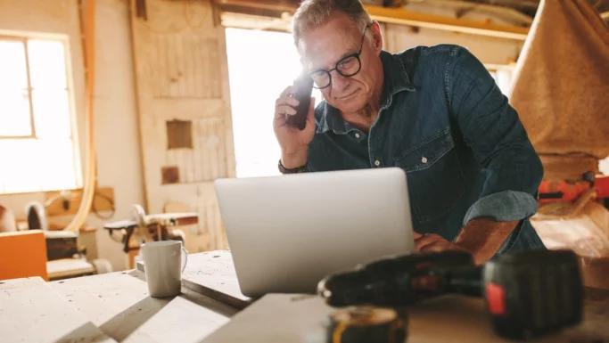 En ældre mand med briller og en denimskjorte taler i telefon, mens han arbejder på en bærbar computer i et værksted. På træbordet foran ham står en krus og elværktøj, mens sollyset strømmer ind i arbejdsrummet. Selv som pensionister fortsætter nogle mennesker med at arbejde og sætter dermed nye standarder for engagement og passion inden for deres valgte håndværk.