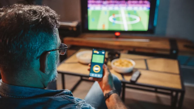 En mand nyder at se en fodboldkamp i tv, mens han tjekker sin smartphone. Der står to skåle med snacks og en fjernbetjening på træbordet foran ham.