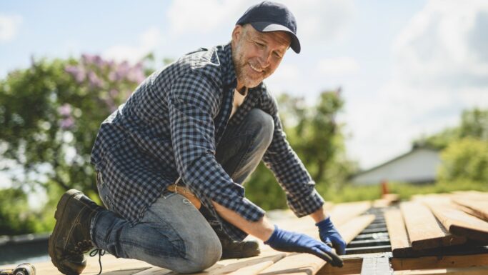 En mand i en ternet skjorte, jeans og handsker sidder på knæ udendørs og arbejder med træplanker, mens han bygger en ny træterrasse. Han smiler og har en kasket på. I den slørede baggrund ses træer og et hus under en delvis overskyet himmel.