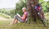 En ældre kvinde sidder barfodet under et træ i en grøn park og nyder sin fritid, mens hun læser en bog. En rød cykel med en kurv står parkeret op ad træet ved siden af hende. Scenen er fredfyldt og omgivet af grønne omgivelser, hvilket indbyder til stille fordybelse og undersøgelse af verden omkring hende.