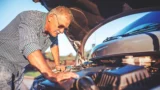 En ældre mand iført solbriller og en ternet skjorte undersøger motoren på en bil med åbent motorhjelm på en solrig dag, muligvis på jagt efter årsagen til en fejl – en almindelig situation, som mange bilister oplever, inden de ringer efter vejhjælp.