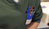 En person i grønne operationsklæder har kuglepenne, en saks og et navneskilt i brystlommen, hvilket tyder på, at vedkommende er sundhedspersonale, sandsynligvis en sygeplejerske eller en læge, i et klinisk miljø – måske en person, der er tilknyttet Medicinrådet eller tager sig af patienter med sjælden hjernekræft.