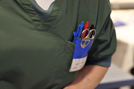En person i grønne operationsklæder har kuglepenne, en saks og et navneskilt i brystlommen, hvilket tyder på, at vedkommende er sundhedspersonale, sandsynligvis en sygeplejerske eller en læge, i et klinisk miljø – måske en person, der er tilknyttet Medicinrådet eller tager sig af patienter med sjælden hjernekræft.