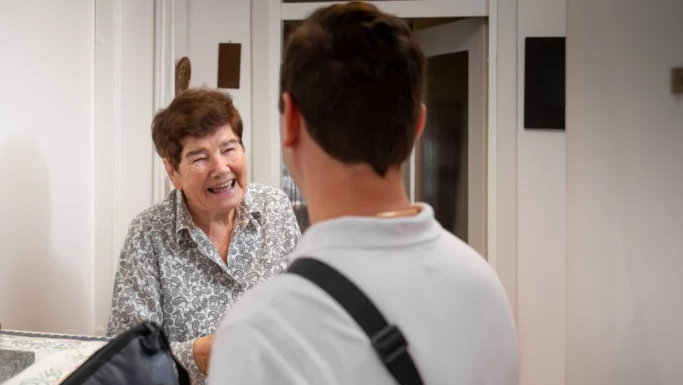 En ældre kvinde smiler, mens hun taler med en yngre mand, der har en taskerem over skulderen, i et lyst rum med hvide vægge og en åben døråbning.
