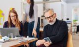 En mand med gråt hår og briller kigger på sin telefon, mens to kvinder – den ene siddende ved en bærbar computer og den anden stående – arbejder sammen ved et rodet skrivebord i et lyst, moderne kontor – hvilket illustrerer den aktive rolle, som seniorer i 50’erne stadig kan spille på nutidens dynamiske arbejdspladser.