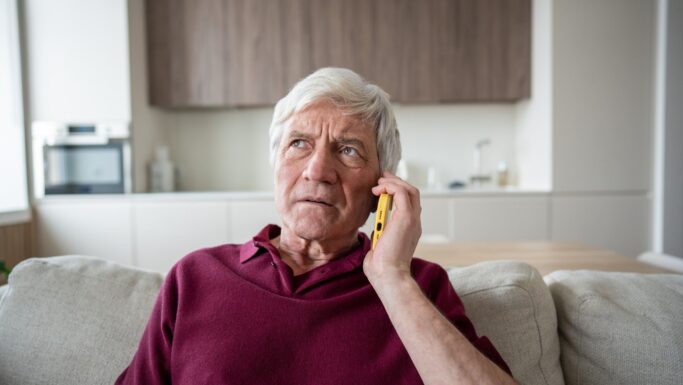En ældre mand med gråt hår, iført en rødbrun skjorte, sidder på en sofa med en gul telefon mod øret; han ser bekymret ud. Han virker fuldstændig opslugt af samtalen, som om han venter på et vigtigt svar. »Hører du?« spørger han måske ængsteligt ind i telefonrøret, i håb om at få en beroligende besked fra den anden ende. I baggrunden bidrager et moderne køkken til scenens nutidige stemning. Rummet er godt oplyst og pænt indrettet, hvilket tyder på en velordnet livsstil. Trods sin ro er der bekymringsrynker indgraveret i hans ansigt, mens han lytter opmærksomt gennem røret. Dette øjeblik indfanger både spænding og forventning, mens han afventer et svar, der kan berolige ham (7 sætninger).