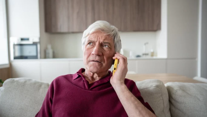 En ældre mand med gråt hår, iført en rødbrun skjorte, sidder på en sofa med en gul telefon mod øret; han ser bekymret ud. Han virker fuldstændig opslugt af samtalen, som om han venter på et vigtigt svar. »Hører du?« spørger han måske ængsteligt ind i telefonrøret, i håb om at få en beroligende besked fra den anden ende. I baggrunden bidrager et moderne køkken til scenens nutidige stemning. Rummet er godt oplyst og pænt indrettet, hvilket tyder på en velordnet livsstil. Trods sin ro er der bekymringsrynker indgraveret i hans ansigt, mens han lytter opmærksomt gennem røret. Dette øjeblik indfanger både spænding og forventning, mens han afventer et svar, der kan berolige ham (7 sætninger).
