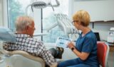 En tandlæge i blå kittel viser en ældre mand røntgenbilleder af tænderne på en tablet i en lys, moderne tandlægeklinik, mens de taler om det nye initiativ med gratis tandlægehjælp, som ministeren har annonceret til fordel for 77.000 ældre.