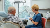 En tandlæge i blå kittel viser en ældre mand røntgenbilleder af tænderne på en tablet i en lys, moderne tandlægeklinik, mens de taler om det nye initiativ med gratis tandlægehjælp, som ministeren har annonceret til fordel for 77.000 ældre.