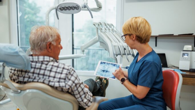 En tandlæge i blå kittel viser en ældre mand røntgenbilleder af tænderne på en tablet i en lys, moderne tandlægeklinik, mens de taler om det nye initiativ med gratis tandlægehjælp, som ministeren har annonceret til fordel for 77.000 ældre.