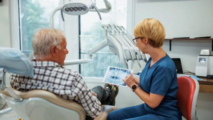 En tandlæge i blå kittel viser en ældre mand røntgenbilleder af tænderne på en tablet i en lys, moderne tandlægeklinik, mens de taler om det nye initiativ med gratis tandlægehjælp, som ministeren har annonceret til fordel for 77.000 ældre.