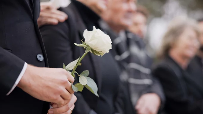 En person i sort tøj holder en enkelt hvid rose i hånden og er omgivet af andre i mørkt tøj, hvilket tyder på en højtidelig eller mindehøjtidelighed for ægtefæller, der har været udsat for den samme ulykke.