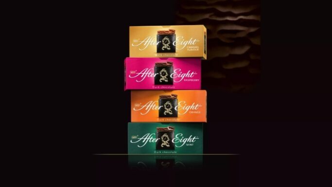 Fire »After Eight«-chokoladeæsker stablet lodret i smagsvarianterne karamel, hindbær, appelsin og mynte, hvor hver enkelt byder på en unik smagsoplevelse af chokolade, fremhævet på en mørk baggrund med en blød skygge ovenover.