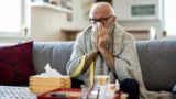 En ældre mand, indpakket i et tæppe, sidder på en sofa og pudser næsen med et papirlommetørklæde. På bordet foran ham står en æske papirlommetørklæder og forskellige lægemidler, herunder allergimedicin, hvilket tyder på, at han måske er ramt af en forkølelse eller influenza.