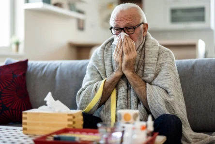 En ældre mand, indpakket i et tæppe, sidder på en sofa og pudser næsen med et papirlommetørklæde. På bordet foran ham står en æske papirlommetørklæder og forskellige lægemidler, herunder allergimedicin, hvilket tyder på, at han måske er ramt af en forkølelse eller influenza.