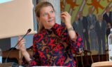 En kvinde i en farverig, mønstret kjole taler ved et talerstol med en mikrofon, muligvis til seniorer og arbejdsgivere; i baggrunden ses et vægmaleri med mennesker i jakkesæt, hvilket tyder på, at der er indflydelsesrige personer til stede, såsom en minister.