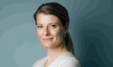 En kvinde med lysebrunt hår, der er sat op, smiler blidt ind i kameraet. Hun er iført en lys bluse og små guldøreringe og står foran en dæmpet blågrå baggrund – hvilket afspejler en stille optimisme, efterhånden som nyheden om, at pensionen stiger fra 1. januar, blandt andet med Arne-pensionen, breder sig i samfundet.