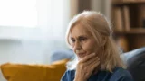 En ældre kvinde med lyst hår sidder indenfor, med hånden på hagen og et eftertænksomt udtryk i ansigtet – måske tænker hun over de forandringer, der følger med overgangsalderen. Blødt lys strømmer ind gennem et vindue i nærheden, og i baggrunden ses en bogreol og en gul pude, hvilket skaber en fredfyldt stemning, som mange kvinder måske søger efter en lang dag på arbejde.