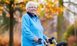 En ældre kvinde med hvidt hår, der bærer briller og en lyseblå jakke, står udendørs i Danmark med en rollator i hånden. I baggrunden ses efterårstræer med gule og orange blade, der fremstår slørede. Hun smiler og er et levende eksempel på fordelene ved sundhed i alle aldre.