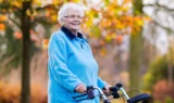 En ældre kvinde med hvidt hår, der bærer briller og en lyseblå jakke, står udendørs i Danmark med en rollator i hånden. I baggrunden ses efterårstræer med gule og orange blade, der fremstår slørede. Hun smiler og er et levende eksempel på fordelene ved sundhed i alle aldre.