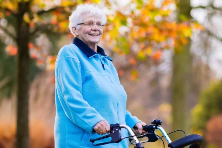 En ældre kvinde med hvidt hår, der bærer briller og en lyseblå jakke, står udendørs i Danmark med en rollator i hånden. I baggrunden ses efterårstræer med gule og orange blade, der fremstår slørede. Hun smiler og er et levende eksempel på fordelene ved sundhed i alle aldre.