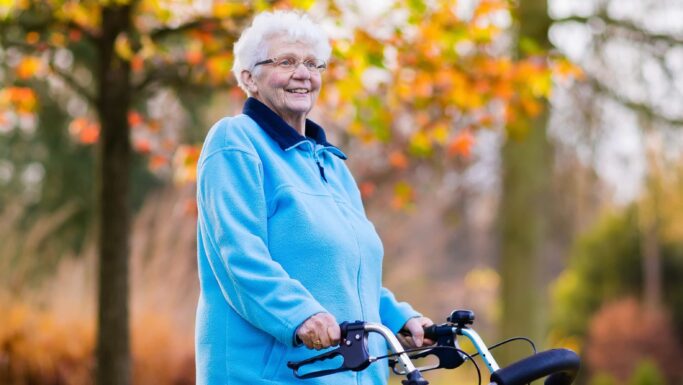 En ældre kvinde med hvidt hår, der bærer briller og en lyseblå jakke, står udendørs i Danmark med en rollator i hånden. I baggrunden ses efterårstræer med gule og orange blade, der fremstår slørede. Hun smiler og er et levende eksempel på fordelene ved sundhed i alle aldre.