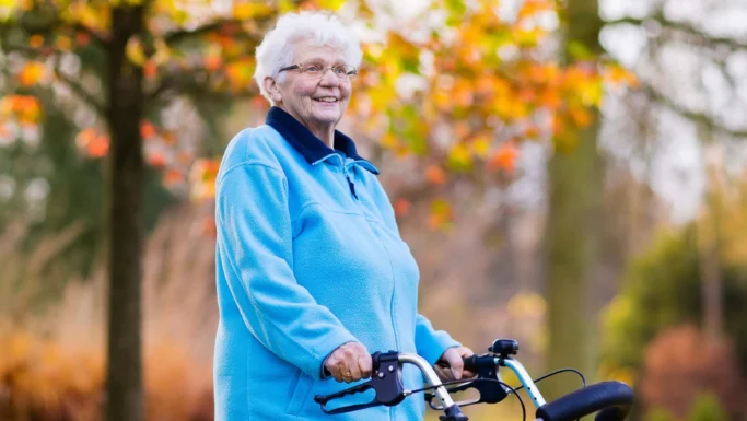 En ældre kvinde med hvidt hår, der bærer briller og en lyseblå jakke, står udendørs i Danmark med en rollator i hånden. I baggrunden ses efterårstræer med gule og orange blade, der fremstår slørede. Hun smiler og er et levende eksempel på fordelene ved sundhed i alle aldre.