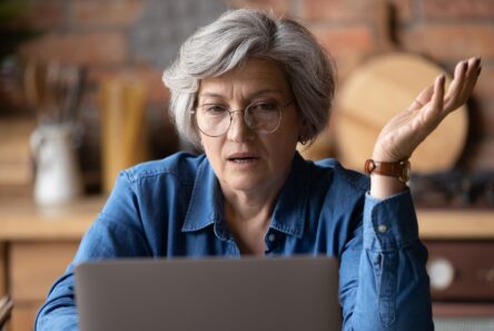 En ældre kvinde med gråt hår og briller ser forvirret eller frustreret ud, mens hun sidder ved et bord med en bærbar computer og løfter den ene hånd i en spørgende gestus. Scenen indfanger de bekymringer, som mange ældre oplever, når de skal håndtere økonomi, idet baggrunden viser et hyggeligt køkken med en murstensvæg og køkkenredskaber af træ.