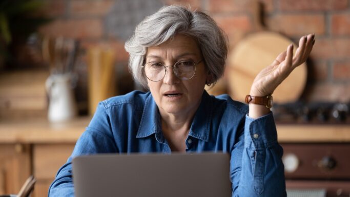 En ældre kvinde med gråt hår og briller ser forvirret eller frustreret ud, mens hun sidder ved et bord med en bærbar computer og løfter den ene hånd i en spørgende gestus. Scenen indfanger de bekymringer, som mange ældre oplever, når de skal håndtere økonomi, idet baggrunden viser et hyggeligt køkken med en murstensvæg og køkkenredskaber af træ.