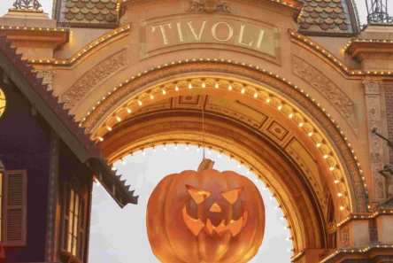 En stor jack-o-lantern-dekoration hænger under den oplyste Halloween-Tivoli-buegang, med lys i efterårsmotiver og en festlig Halloween-stemning – perfekt til at indfange magien, inden du tager din billetter eller turpas med på et uforglemmeligt besøg.