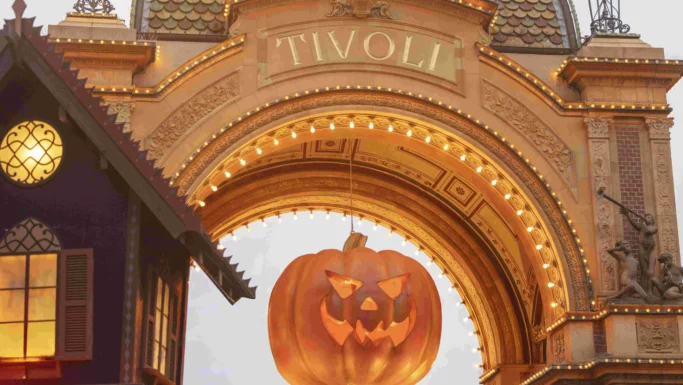 En stor jack-o-lantern-dekoration hænger under den oplyste Halloween-Tivoli-buegang, med lys i efterårsmotiver og en festlig Halloween-stemning – perfekt til at indfange magien, inden du tager din billetter eller turpas med på et uforglemmeligt besøg.