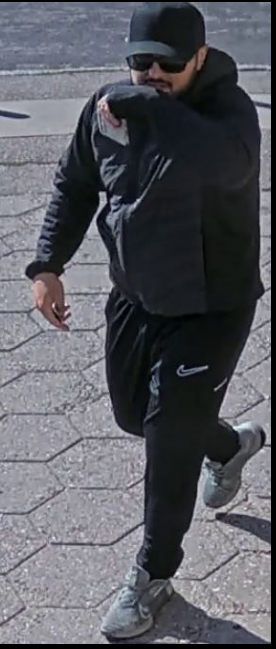 En person iført sort kasket, solbriller, sort jakke, sorte Nike-bukser og grå sneakers går på fortovet, mens vedkommende dækker munden med ærmet – et billede, der er offentliggjort, da politiet efterlyser en mand i forbindelse med syv bedragerisager.