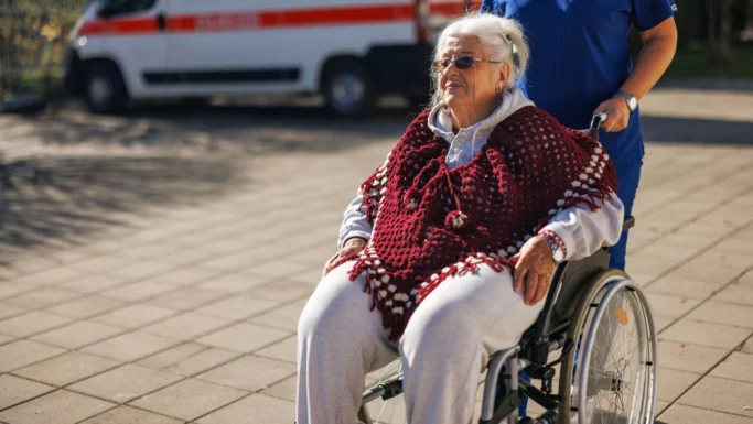 En ældre kvinde iført solbriller og et rødt sjal sidder i en kørestol, der skubbes af en plejer udenfor, med en ambulance i baggrunden; hun venter muligvis på flextrafik-transport på grund af de stramme regler, der er indført af regeringspartiet.