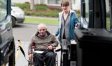 En ældre mand i kørestol får hjælp af en kvinde, mens han stiger om bord på et Flextrafik-køretøj ved hjælp af en kørestolslift. De befinder sig udendørs i nærheden af en parkeret varevogn og er muligvis på vej til eller fra sygehuset.