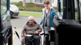 En ældre mand i kørestol får hjælp af en kvinde, mens han stiger om bord på et Flextrafik-køretøj ved hjælp af en kørestolslift. De befinder sig udendørs i nærheden af en parkeret varevogn og er muligvis på vej til eller fra sygehuset.