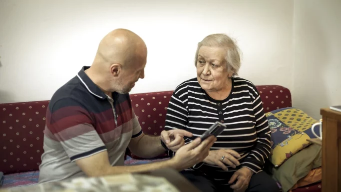 En mand viser en smartphone til en ældre kvinde, der sidder på en rød sofa. Begge ser ud til at være optaget af en samtale, måske om svindel og sikkerhed på nettet. Kvinden har en stribet bluse på, og manden er skaldet og har et kort skæg.
