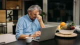 En mand sidder ved et bord med en bærbar computer og holder sig for panden i frustration – måske efter at være blevet kontaktet af svindlere. På bordet står der en kop kaffe og en frugtskål med appelsiner og ananas.
