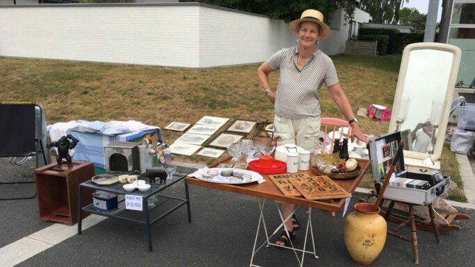 En kvinde med en hat står bag et bord på et loppemarked, hvor der er udstillet vintage-genstande, keramik og spil på borde og på jorden uden for et hus på en overskyet dag – et indbydende syn, der inspirerer dig til at rydde op i dit eget hjem og omfavne et lettere liv ved at opdage unikke skatte.