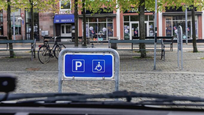Et blåt parkeringsskilt med et kørestolssymbol angiver en reserveret handicapplads – hvor der kræves et gyldigt handicapkort – på en brostensbelagt gade med bænke, træer og et hotel i baggrunden. Sådanne afmærkede områder kontrolleres regelmæssigt af politiet for at afsløre snyd og sikre retfærdig adgang for dem, der har mest brug for det.