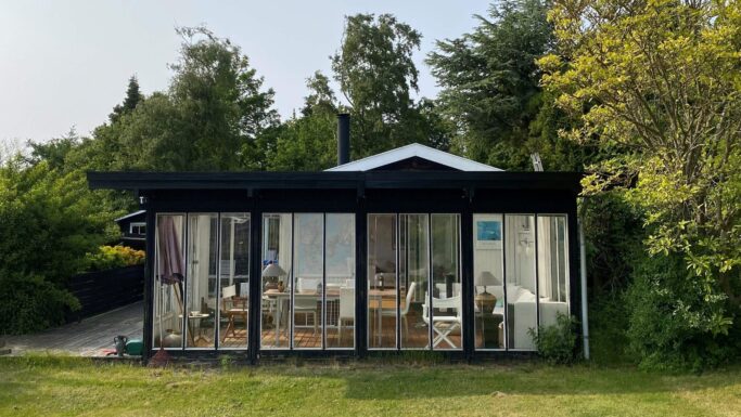 En moderne sort hytte med vinduer fra gulv til loft ligger midt i frodig grønne omgivelser og afslører et hyggeligt, solfyldt interiør med trægulve, stole og lamper. Omgivet af træer og græs indbyder de fredfyldte omgivelser til stille fordybelse – måske endda over emner som privatøkonomi eller overvejelser om fordelene ved et afdragsfrit lån.